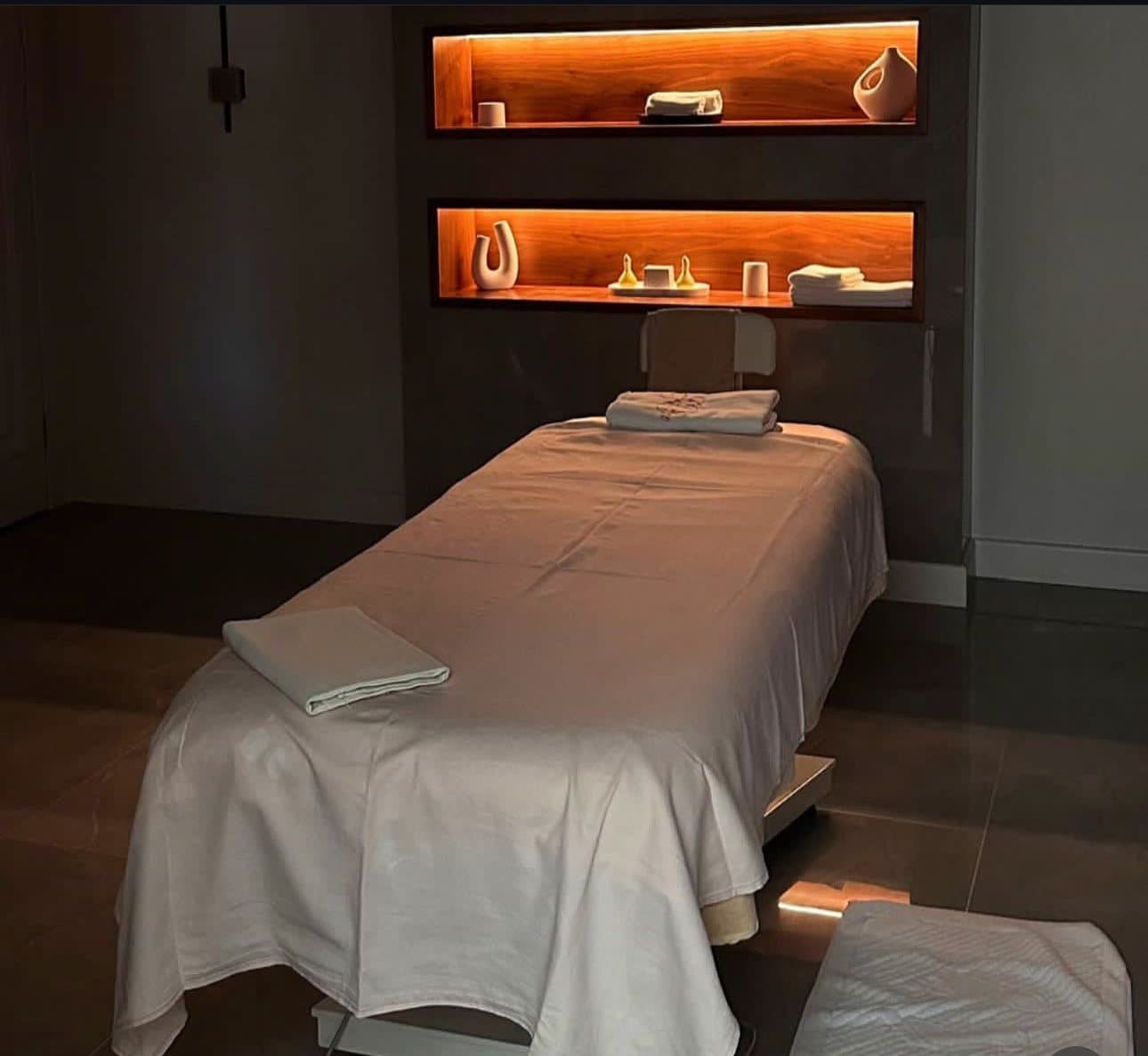 Massage table with white linens in a quiet Miami studio, warm lit shelves behind